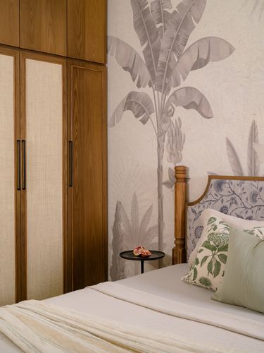 Bedroom corner with cane-panelled wardrobe, banana leaf mural wall, and upholstered wood bedframe with printed cushions