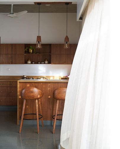 Wooden kitchen island with two curved stools, pendant lights, and a sheer curtain in foreground