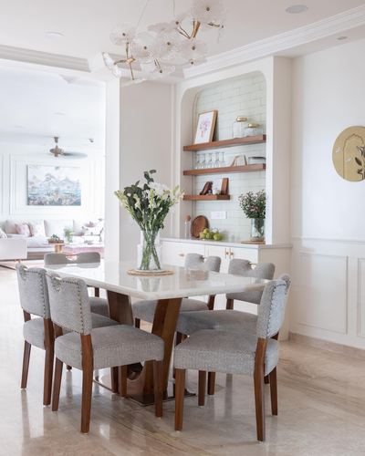 White marble dining table with grey upholstered chairs beneath a glass chandelier, arched tiled niche behind
