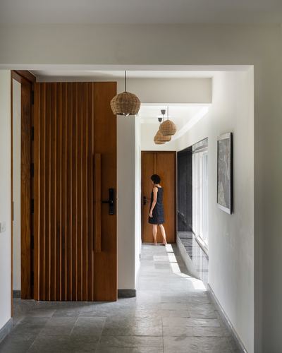 Woman in dark dress stands in a hallway with wood door and rattan pendants, Kochi home