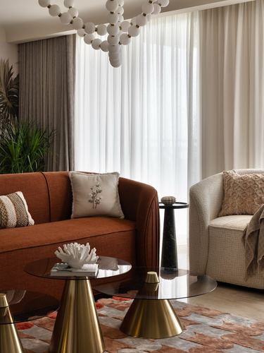 Living room corner with rust curved sofa, glass-brass coffee tables, and sheer-curtained window under a bubble chandelier