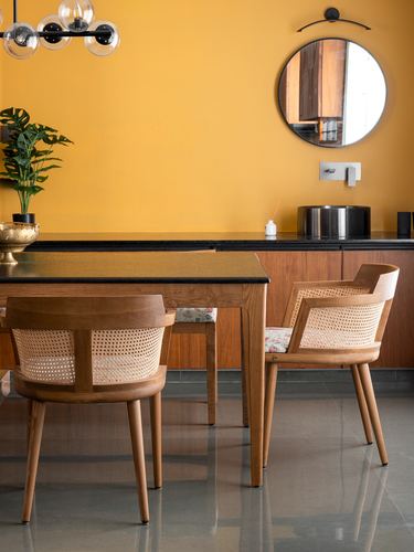 Dining nook with cane-back wooden chairs, black-topped table, and round mirror against mustard wall