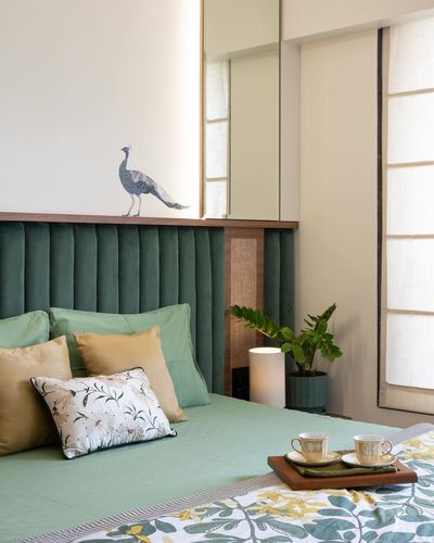 Bedroom with green channel-tufted headboard, mustard and floral pillows, peacock decal, and morning tea tray on bed.