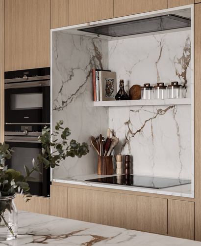 Kitchen niche with veined stone backsplash, wood cabinetry, built-in ovens and open shelf styling.