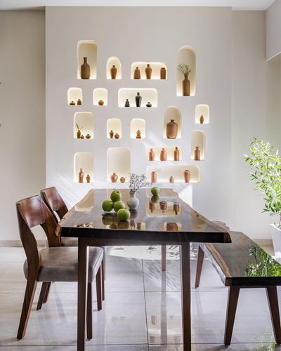 Dining table under wall of backlit niche shelves filled with ceramic vases in Vadodara home