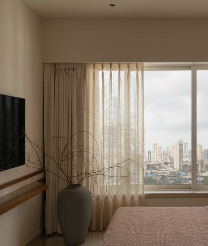 Bedroom corner with sheer curtains, large vase of dried branches, and Mumbai skyline through window