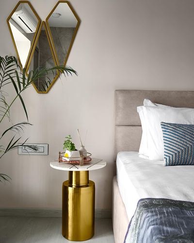 Bedroom corner with brass-framed hexagonal mirror, marble-top brass side table, and beige upholstered bed
