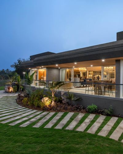 Twilight exterior of modern house with glass facade, chandeliers glowing inside, and linear stone garden path