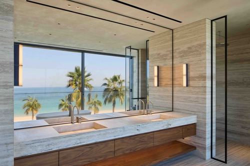 Stone bathroom vanity with wood cabinetry facing floor-to-ceiling window overlooking beach and palm trees