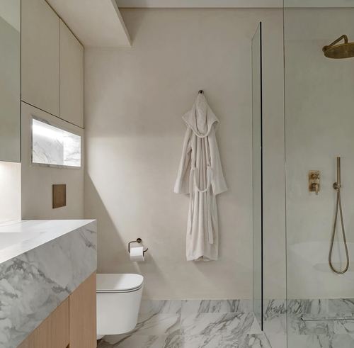 Minimalist marble bathroom with wall-hung toilet, bathrobe on hook, and glass-enclosed shower with brass fixtures