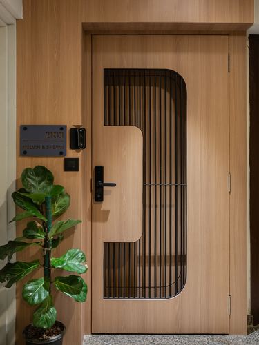 Wooden entrance door with curved black metal grille, nameplate, and fiddle leaf fig plant beside it