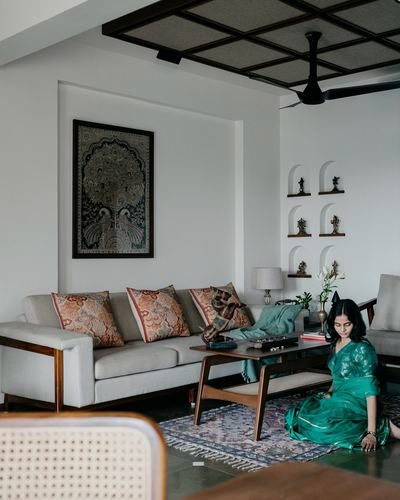Woman in green saree sits on floor beside wooden coffee table in Chennai living room with cane bench and arched niches