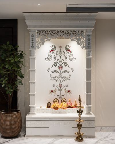 White carved marble pooja unit with peacock inlay and brass diya lamp beside a potted plant