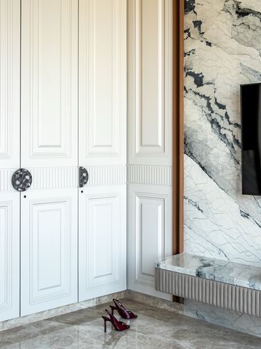 White fluted wardrobe beside marble-clad wall with mounted tv and bench, red heels on floor