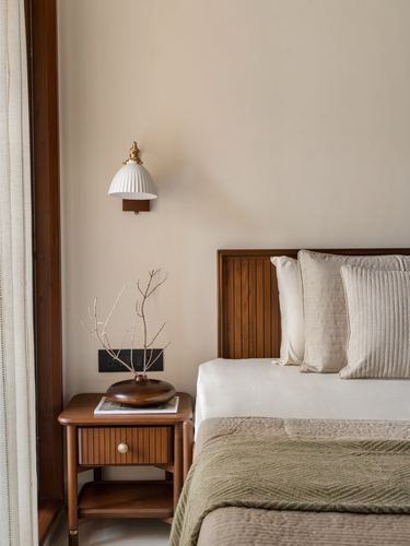 Bedroom corner with fluted ceramic sconce, wooden nightstand, and teak headboard with layered pillows