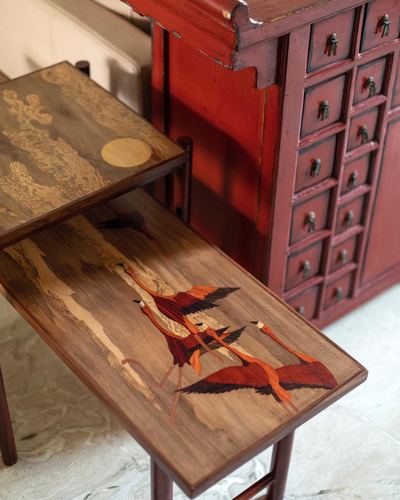 Wooden nesting tables with flamingo marquetry beside a red Chinese medicine cabinet on marble floor