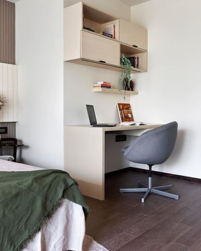 Beige wood study nook with floating desk, wall cabinets, and grey swivel chair beside a bed