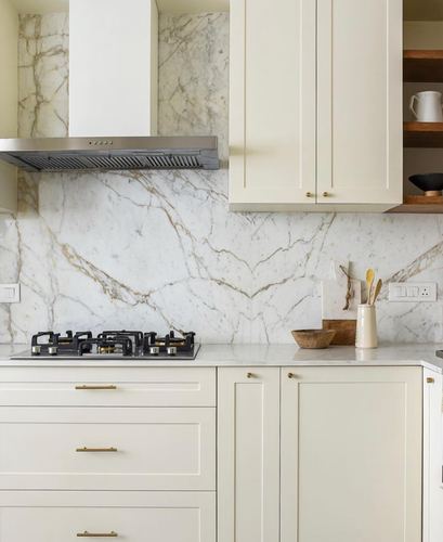 White kitchen with marble backsplash, stainless steel chimney hood, black cooktop, and brass cabinet handles