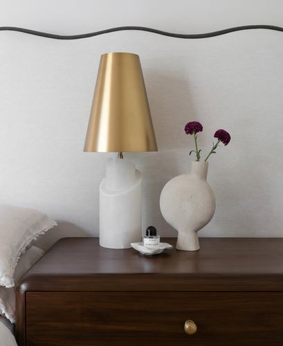 Bedside table with brass-shaded lamp, stone vase with dark flowers, on a dark wood nightstand