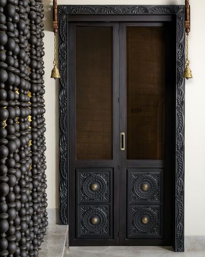 Dark carved wooden double door with glass panels and brass bells beside a sculptural bead-textured wall