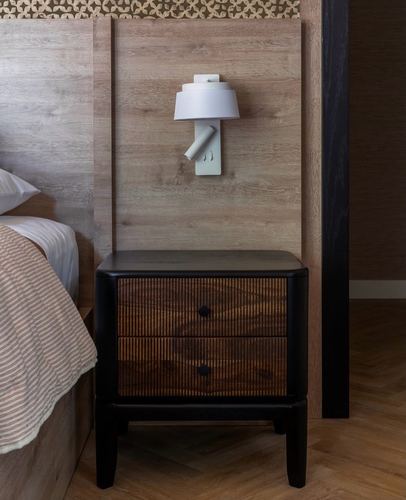 Black nightstand with fluted drawers beside bed under a wall-mounted reading sconce
