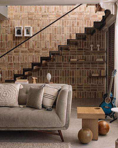 Living room with tiled staircase wall, grey velvet sofa, guitar and sculptural wood decor