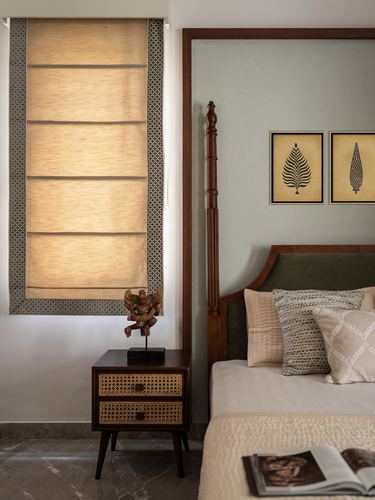 Bedroom corner with carved four-poster bed, cane bedside table, and textured roman blind window