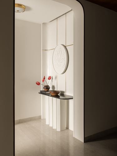 Curved white foyer with fluted panel, circular Ganesha relief, and dark stone console table with red flowers