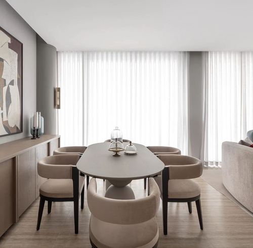 Oval dining table with beige upholstered chairs before sheer curtains in a greige-toned dining room