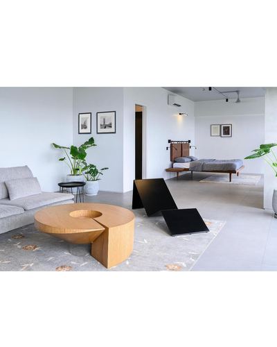Open-plan minimalist studio with sculptural wood coffee table, grey sofa, and floating bed beyond