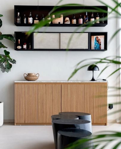Fluted wood sideboard beneath black floating shelves styled with bottles and art, framed by plants