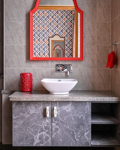 Bathroom vanity with white vessel sink, grey marble cabinet, and red arched mirror reflecting patterned tile wall