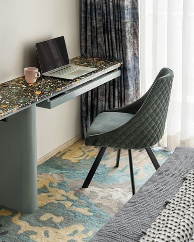 Terrazzo desk with laptop and mug beside a green quilted chair on a patterned rug