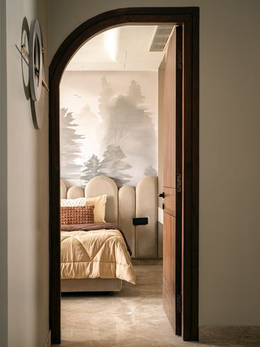View through an arched wooden door into a bedroom with scalloped headboard and forest mural