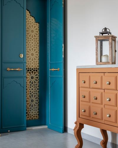 Teal arched cupboard doors open to reveal a golden jali screen beside a terracotta drawer cabinet with lantern