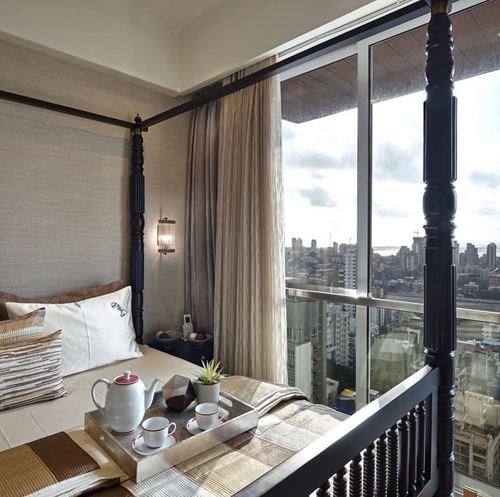Four-poster dark wood bed with breakfast tray beside a floor-to-ceiling window overlooking a city skyline