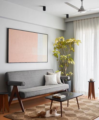 Grey mid-century sofa below a pink abstract painting, plant beside sheer curtained window, cat on jute rug