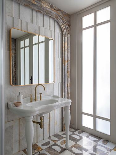 Marble-clad bathroom with arched niche, brass-framed mirror, and console sink beside tall window