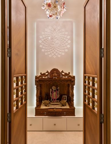 Symmetrical carved wooden pooja mandir framed by open doors with brass bells, backlit om motif above
