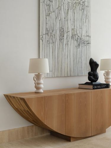 Wood sideboard with curved base under abstract artwork, flanked by ceramic lamps and a black sculpture.