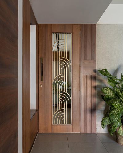 Wooden entrance door with brass arch pattern glass panel beside fluted wood wall and plant