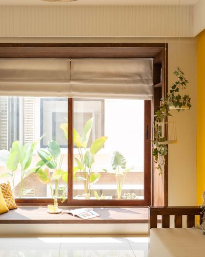Wooden window seat with cushions beside tall glass window and hanging plants, yellow wall accent