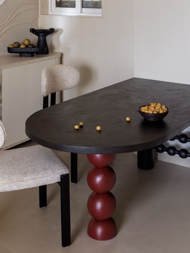 Oval dark stone dining table with red stacked-sphere leg, boucle chairs, fruit bowl on top