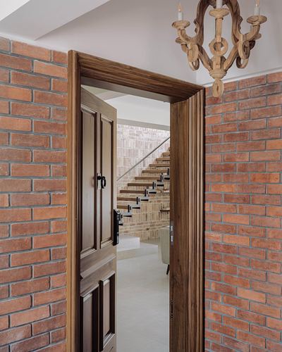 Open wood door framed by exposed brick walls reveals a staircase and hanging carved chandelier beyond.