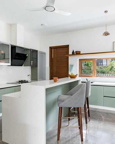 Sage-green kitchen with curved white island, two grey bar stools, wooden door in background