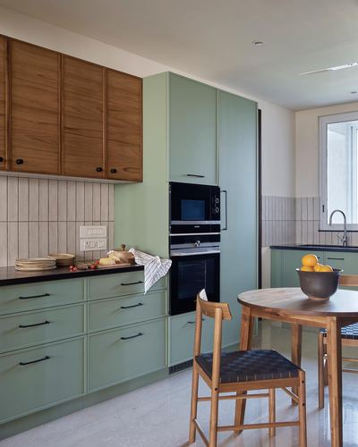 Sage green kitchen with wood upper cabinets, built-in ovens, and a round wood dining table with oranges
