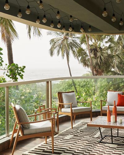 Curved balcony with teak armchairs, checkered rug, and coffee table overlooking palm trees and the sea in Mumbai.