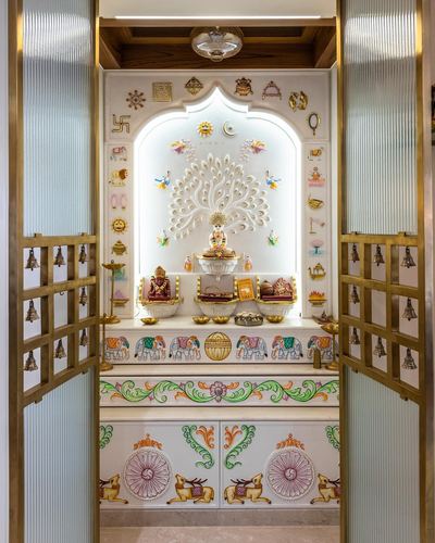 Ornate white marble pooja unit with carved peacock, brass bells and hand-painted elephant motifs, seen through open reeded glass doors.