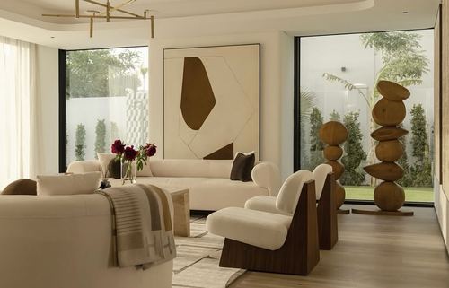 Cream living room with wood-backed armchairs, abstract art, and stacked wood sculptures by garden windows