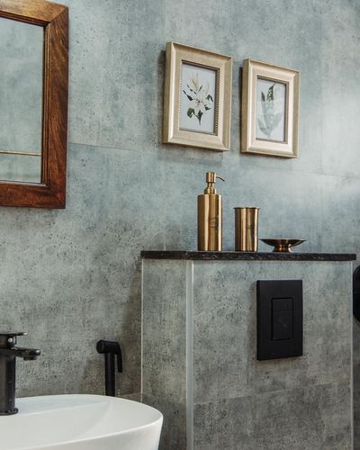 Bathroom corner with grey stone walls, brass accessories, framed botanical prints, and wood mirror frame